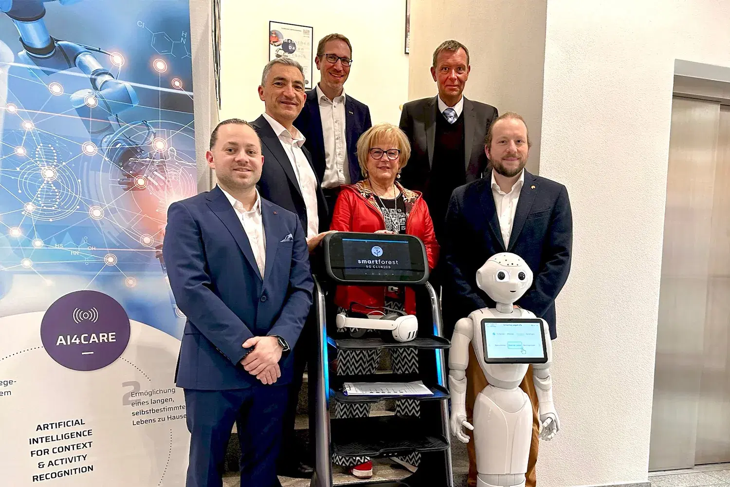 階段に並ぶ6人の集合写真。手前に、ディスプレイに SMART FOREST 5G Clinics のロゴを表示した配送ロボットと、タブレットを備えた人型ロボット